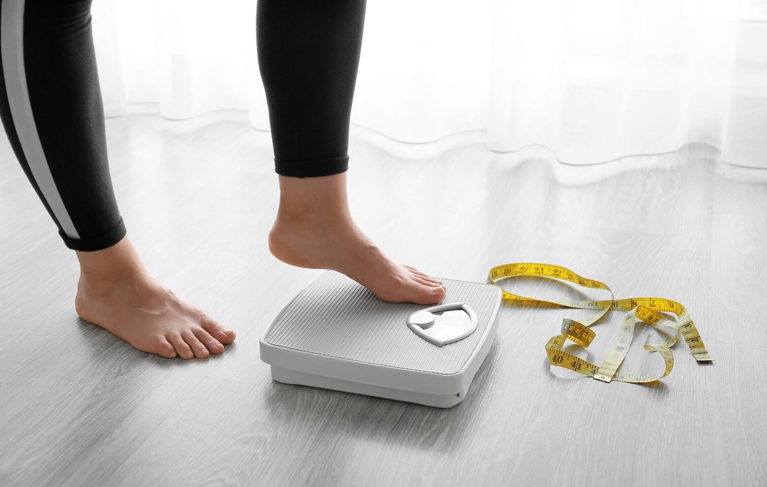 Τα πόδια μιας γυναίκας καθώς ανεβαίνει στη ζυγαριά, ενώ δίπλα της στο πάτωμα υπάρχει και μια μεζούρα μέτρησης.