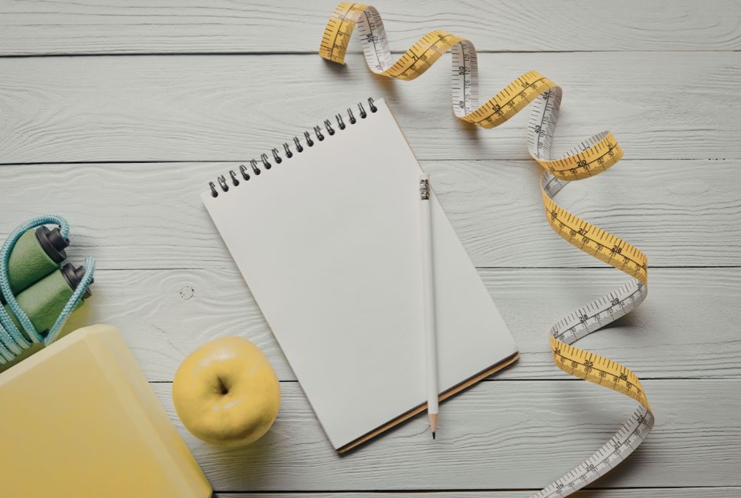 Πάνω σε μια ξύλινη επιφάνεια ένα σημειωματάριο με ένα στύλο και δίπλα υπάρχει μια μεζούρα μέτρηση, ένα μήλο, και ένα λάστιχο γυμναστικής.